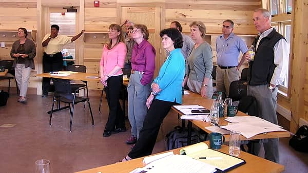 People standing around tables in a conference room, facing a presenter who is out of the frame