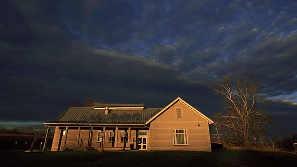 A house standing alone at dusk, its windows lit against a darkening sky and the yard around it in shadow