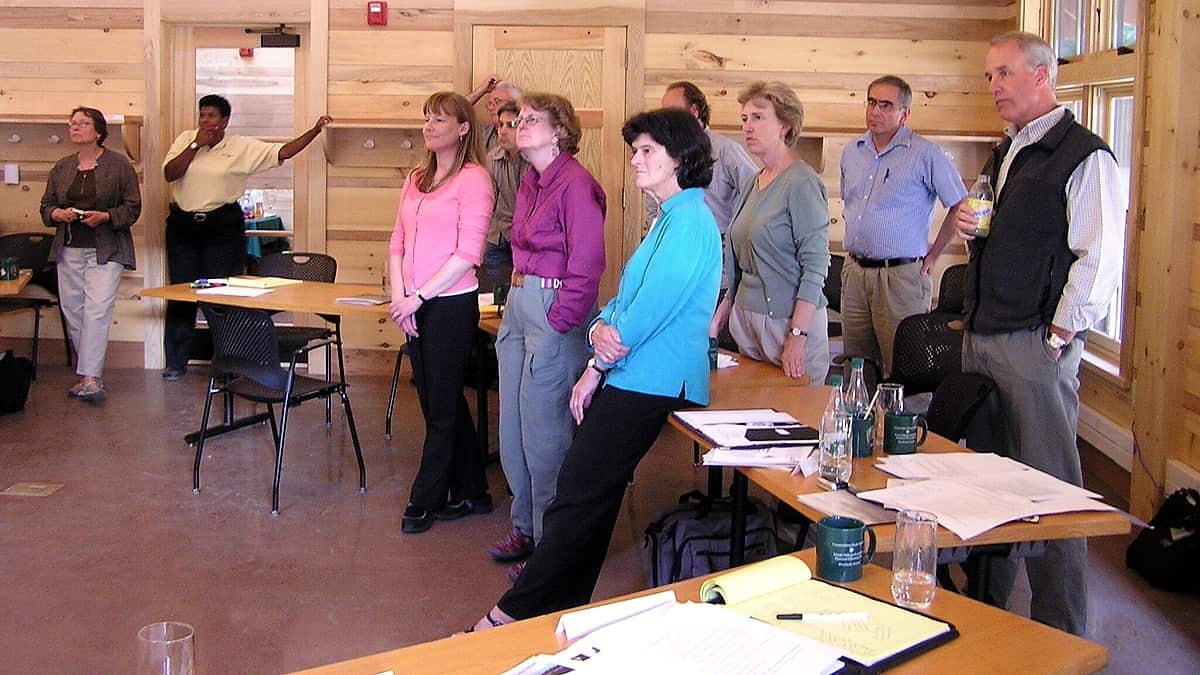 People standing around tables in a conference room, facing a presenter who is out of the frame